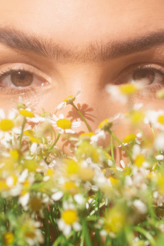A close up of a woman's brown eyes behind a collection of yellow and white daisies.